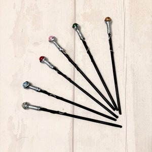 May include: Six black wands with silver tips and colorful gemstone accents. The wands are arranged in a fan shape on a white wooden surface.