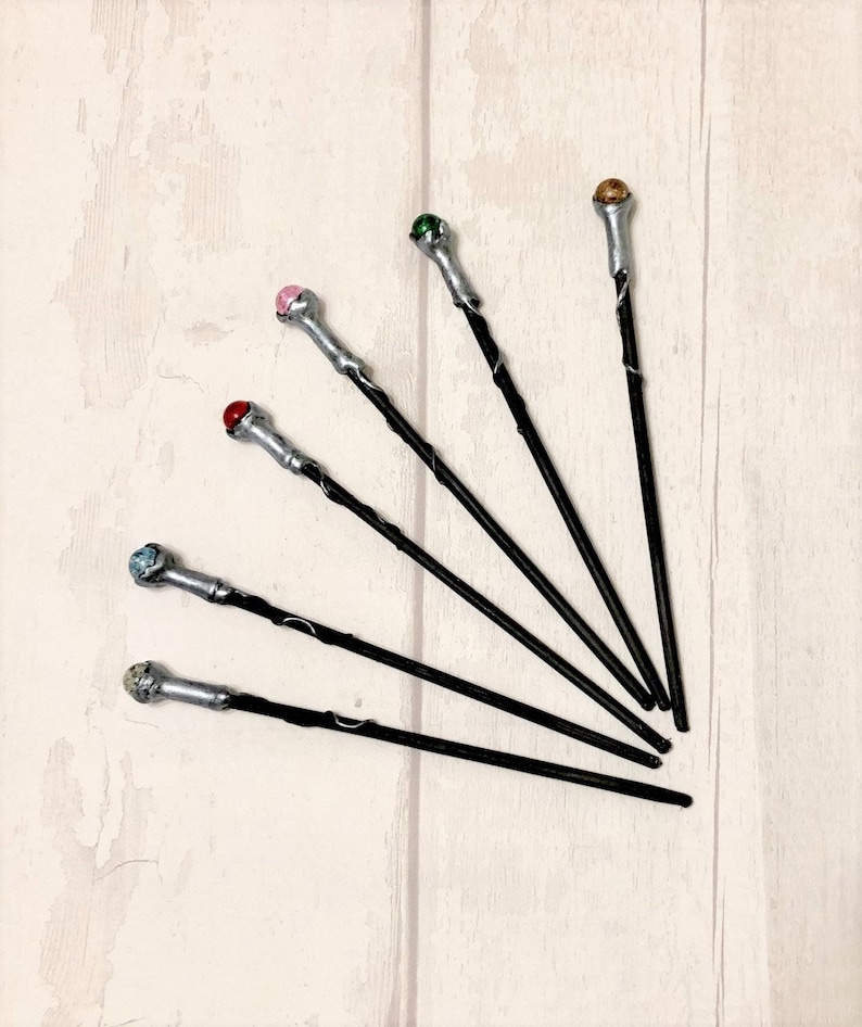 May include: Six black wands with silver tips and colorful gemstone accents. The wands are arranged in a fan shape on a white wooden surface.