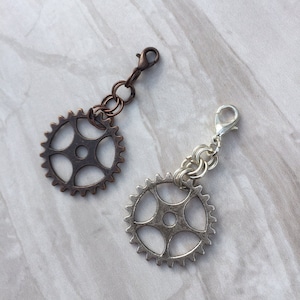May include: Two metal gear charms, one silver and one copper, with a chain and clasp. The charms are on a white marble background.
