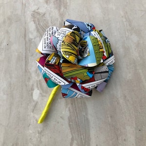 May include: A yellow stem with a flower made from comic book pages. The flower is blue, red, yellow, and green. The comic book pages have text on them, including "NO GOOD! THE WAY", "LOCK HAS KICKED", "IN ALI GOT WAS", "HAVE ENGAGE", "BE LA", "LITTLE TOY OF", "FASCINATE", "YOU M", "AKS THE TRU", and "GOODI".