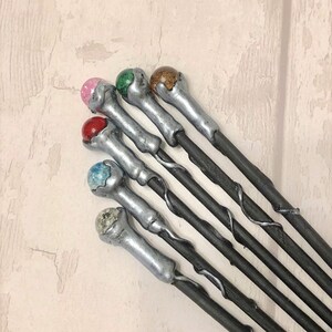 May include: A set of six silver and black wands with different colored gemstone tips. The tips are pink, green, brown, red, blue, and white.