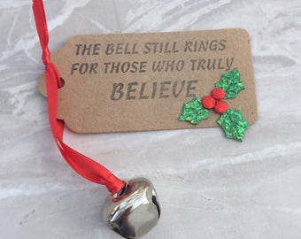 May include: A brown paper tag with the text "The bell still rings for those who truly believe" and a green holly sprig with red berries. A silver bell is attached to a red ribbon.