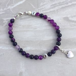 May include: Purple and black agate bead bracelet with silver charm and heart pendant. The bracelet has two silver beads with the numbers 1 and 2 on them.