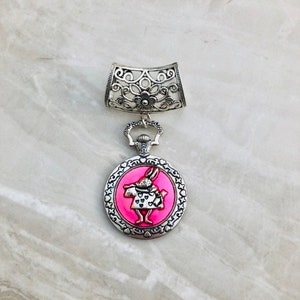May include: Silver pendant with a filigree design and a round charm featuring a pink enamel image of the White Rabbit from Alice in Wonderland.