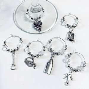 May include: A collection of silver wine glass charms. Each charm features a different wine-related design, including a bunch of grapes, a corkscrew, a bottle, and a wine basket. The charms are adorned with clear beads and silver accents.