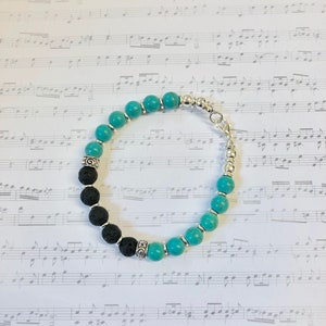 Puede incluir: Un brazalete de cuentas turquesa y negras con un cierre plateado. El brazalete está hecho con cuentas de lava y turquesa.