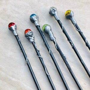May include: Seven silver wands with different colored gemstone tips. The wands have a silver vine design and are arranged in a row.