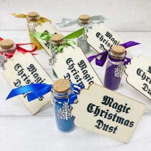 Puede incluir: Una colección de pequeñas botellas de vidrio llenas de purpurina y coronadas con tapones de corcho. Cada botella tiene un encanto de copo de nieve plateado adjunto y una etiqueta de madera que dice "Polvo mágico de Navidad". Las botellas están atadas con cintas azules, rojas, verdes y plateadas.