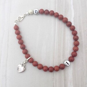 May include: A brown beaded bracelet with silver charms. The charms are a heart and two beads with the letters 'I' and 'J' on them.