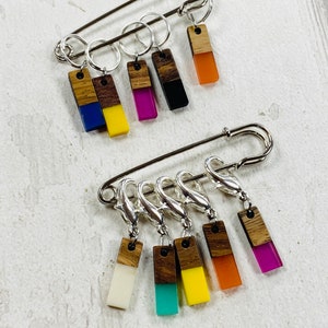 May include: A set of six colorful stitch markers with wooden tops and metal rings. The markers are attached to a silver safety pin.