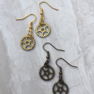 May include: A pair of gold and bronze earrings with small gear charms. The earrings are on a white background.