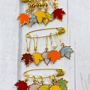May include: A set of nine gold-toned leaf-shaped stitch markers with colourful enamel finishes. The markers are arranged on three safety pins.