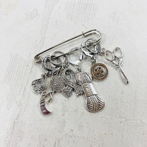 May include: Silver stitch markers with various charms, including a ball of yarn, scissors, and a button. The markers are attached to a safety pin.