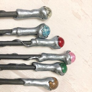 May include: Five silver-painted wooden wands with different colored gemstone tips. The tips are blue, red, pink, green, and brown.