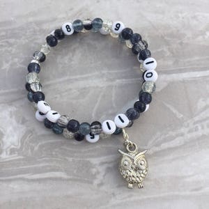 Puede incluir: Un brazalete de cuentas de doble hebra con números negros y blancos y un dije de búho plateado. Las cuentas son transparentes con un patrón moteado gris y negro.