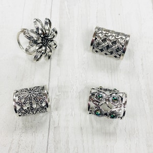 May include: Set of four silver hair cuffs with decorative designs. One cuff features a flower design, another has a woven pattern, and two have floral designs with green gemstones.