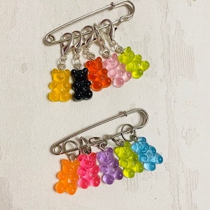 Könnte beinhalten: Ein Satz bunter Gummibärchen-Maschenmarkierer, die an einer silbernen Sicherheitsnadel befestigt sind. Die Markierer sind in den Farben Gelb, Orange, Schwarz, Rosa, Grün, Blau, Lila und Hellblau.