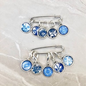 Op de afbeelding: Set van acht blauwe en witte steekmarkeerders met bloemmotieven. De markeerders zijn bevestigd aan een zilveren veiligheidsspeld en een zilveren karabijnhaak.