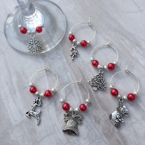 May include: A collection of Christmas-themed wine charms. Each charm features a silver-toned pendant, such as a snowflake, reindeer, or Christmas tree, paired with red beads and silver accents. The charms are designed to attach to the stem of a wine glass.