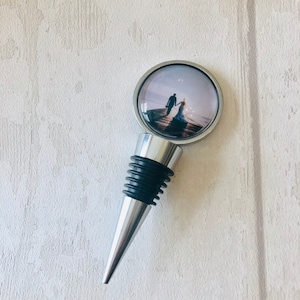May include: Silver bottle stopper with a black rubber seal. The top features a round, clear glass insert with a photo of a couple walking on a beach.