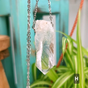 Large Clear Quartz raw crystal necklace with a stainless steel chain shown dangling with plants and crystals in the background.