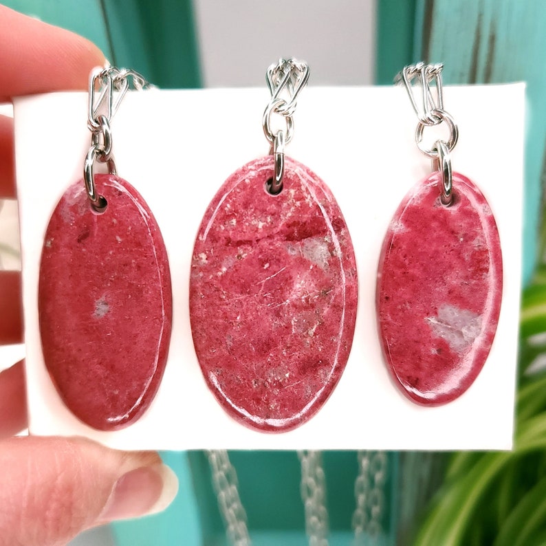 Natural pink polished Thulite stone necklaces with silver tone stainless steel paperclip chains shown against a white background with plants.