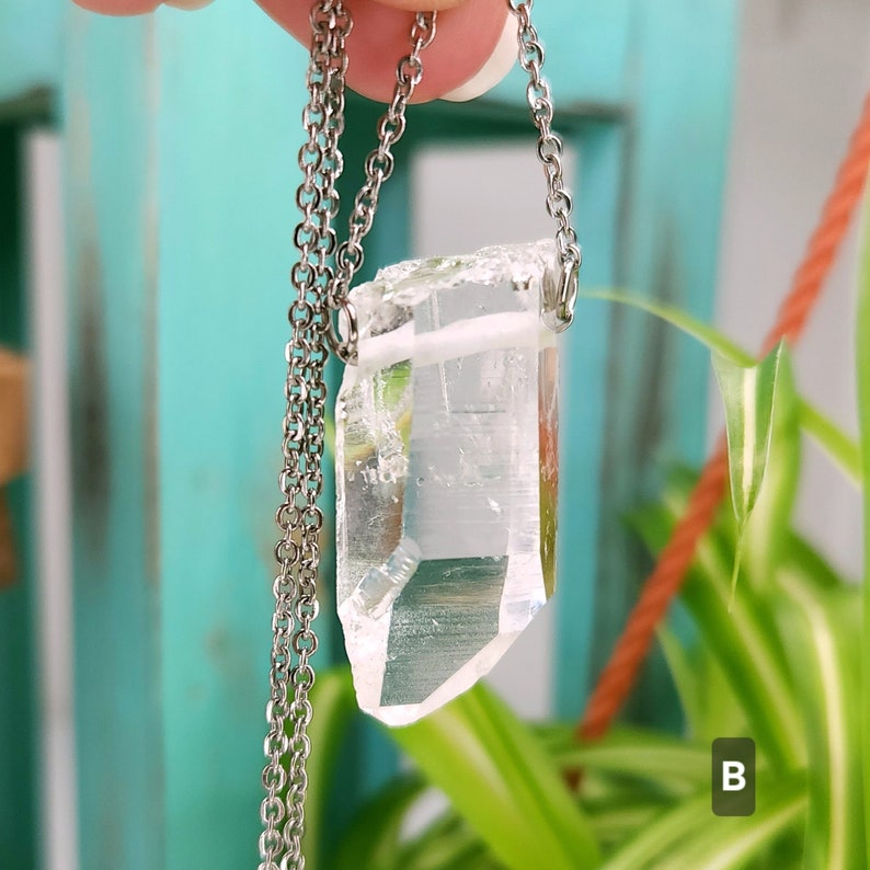 Large Clear Quartz raw crystal necklace with a stainless steel chain shown dangling with plants and crystals in the background.