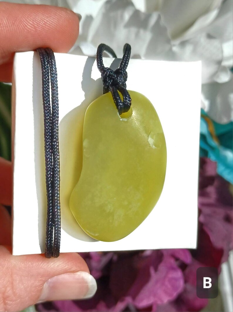 Vibrant green freeform shaped natural Washington Serpentine crystal necklace with an adjustable black cord shown in hand against a white background with flowers and plants.