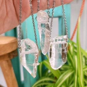 Large Clear Quartz raw crystal necklaces with stainless steel chains shown dangling with plants and crystals in the background.