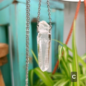 Large Clear Quartz raw crystal necklace with a stainless steel chain shown dangling with plants and crystals in the background.