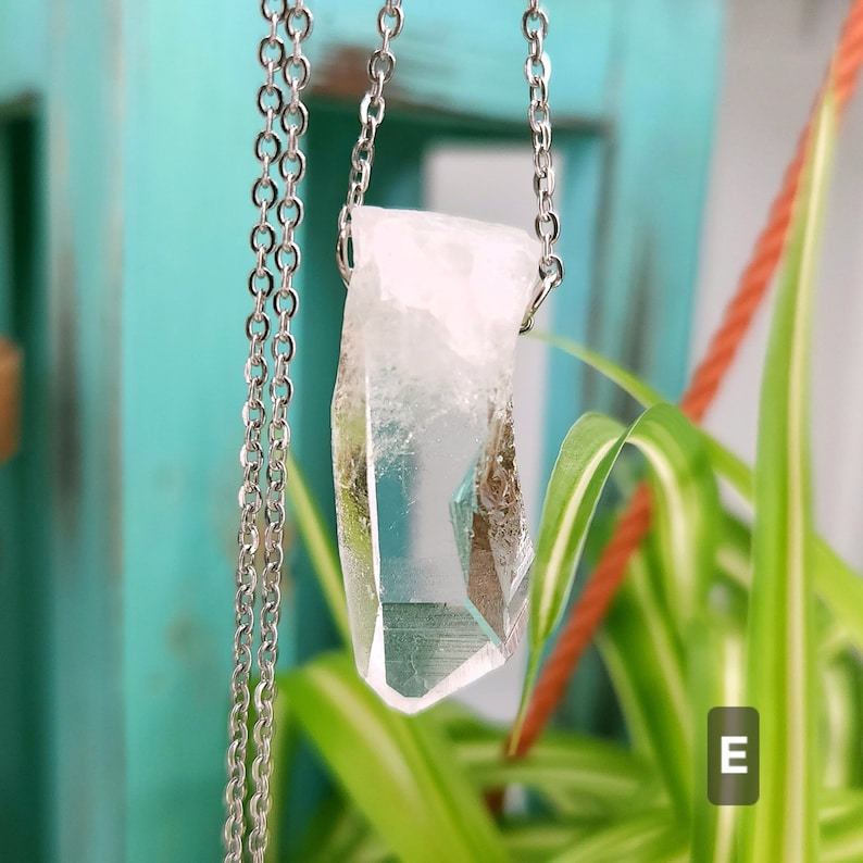 Large Clear Quartz raw crystal necklace with a stainless steel chain shown dangling with plants and crystals in the background.