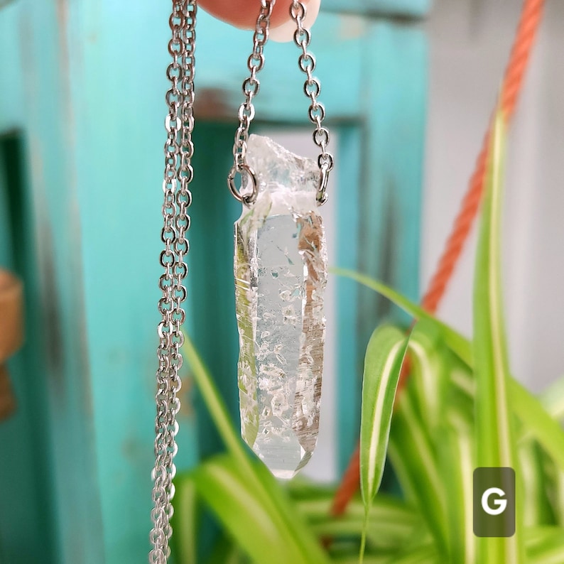 Large Clear Quartz raw crystal necklace with a stainless steel chain shown dangling with plants and crystals in the background.