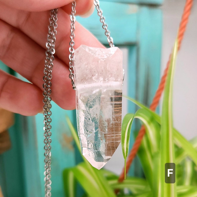 Large Clear Quartz raw crystal necklace with a stainless steel chain shown dangling with plants and crystals in the background.