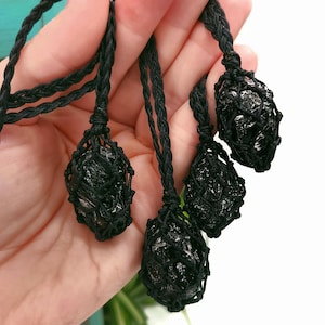 May include: Four black stone pendants are displayed, each encased in a woven black net and suspended from a braided black cord. The stones have a rough, textured appearance. The pendants are held in a hand against a white background.