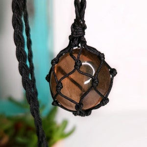 May include: A brown gemstone pendant with a black macrame cord. The gemstone is encased in a black macrame net.