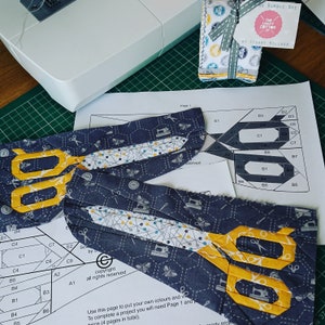 Pode incluir: Um close-up de um padrão de tecido com um design de favo de mel em tons de cinza e branco. O tecido apresenta um padrão repetitivo de máquinas de costura, alfinetes e abelhas. O tecido é cortado na forma de uma tesoura com lâminas amarelas. O texto "The Craft Cotton Co. by Stuart Millard" é visível no canto superior direito da imagem.