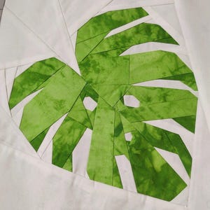 Puede incluir: Una hoja de tela verde sobre un fondo blanco. La hoja está hecha de muchas piezas de tela más pequeñas cosidas juntas para crear un patrón geométrico.