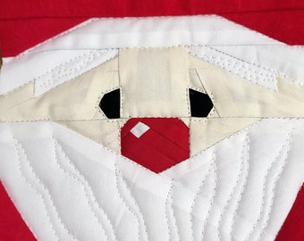May include: Close-up of a quilted Santa Claus face. The design features a white beard and hat, beige skin, black eyes, and a red nose. The background is red, and the stitching is visible.