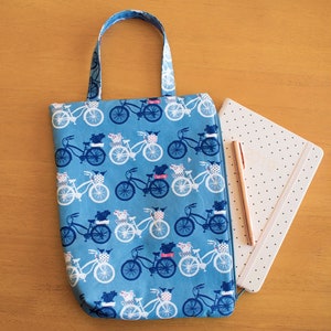 May include: A blue tote bag with a white bicycle pattern. The bag has a zipper closure and a top handle. The bag is sitting on a wooden surface next to a white notebook with gold polka dots.