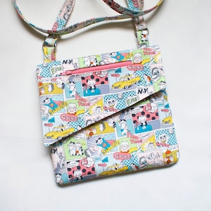 May include: A small crossbody bag with a comic book print featuring cats and the words "Kiss", "Swoosh", "Going", "Yum Chomp", and "Taxi". The bag has a pink zipper and a long adjustable strap.