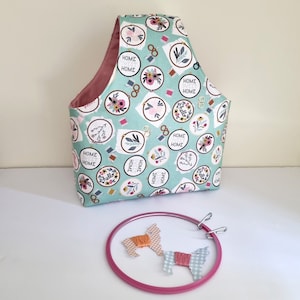 Puede incluir: Una bolsa de proyecto de tela turquesa con un estampado floral y aros de bordado, con las palabras "Home Sweet Home" y "Hello". Un aro de bordado rosa con dos piezas decorativas de hilo y tela está al frente.