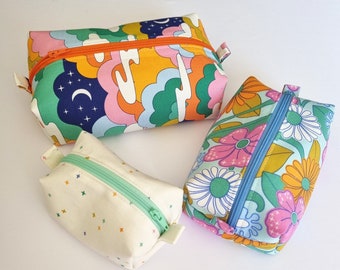 May include: Three colorful zippered pouches with different patterns. The first pouch has a blue, pink, yellow, and green cloud pattern. The second pouch has a white background with a small colorful cross pattern. The third pouch has a blue background with a pink, yellow, and orange floral pattern.