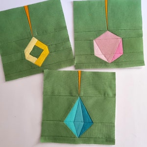 Peut inclure: Trois carrés de tissu vert avec des formes géométriques de couleurs différentes au centre. Les formes sont un diamant jaune, un hexagone rose et un diamant bleu.