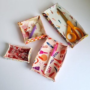 Könnte beinhalten: Vier Stoffaufbewahrungsboxen mit verschiedenen Mustern. Die Boxen sind mit verschiedenen Gegenständen gefüllt, darunter eine Schere, ein rosa Stift und ein Satz bunter Clips.