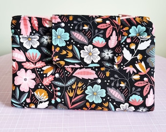 May include: A black fabric wallet with a floral pattern in pink, blue, white, and gold. The wallet has a flap closure and a small pocket on the front.