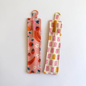 Puede incluir: Dos portalápices de tela. Uno es rosa con un estampado de frutas, fresas, plátanos y flores. El otro es blanco roto con un estampado de ositos de goma rosas y amarillos. Cada uno tiene un bolígrafo dentro y un anillo dorado.