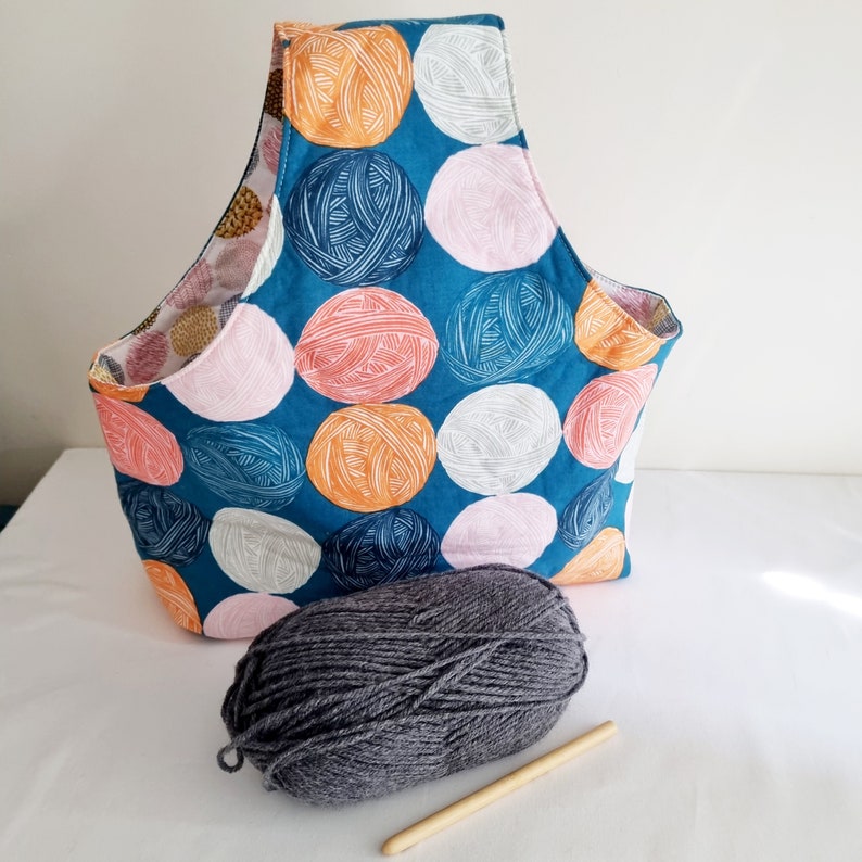 May include: A teal blue fabric tote bag with a handle. The bag is decorated with a pattern of colorful circles. A ball of gray yarn is in front of the bag.