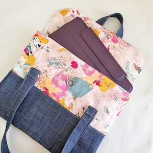 May include: A denim tote bag with a floral fabric lining and a zipped pocket. The bag has a long strap and a smaller pocket on the front. A purple tablet is inside the bag.