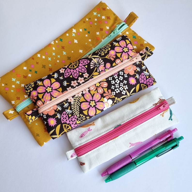 May include: Three fabric pencil cases with different patterns. The top case is yellow with a white crosshatch pattern. The middle case is black with a floral pattern in pink, white, and yellow. The bottom case is white with a pink zipper and a small fish pattern in black and white.