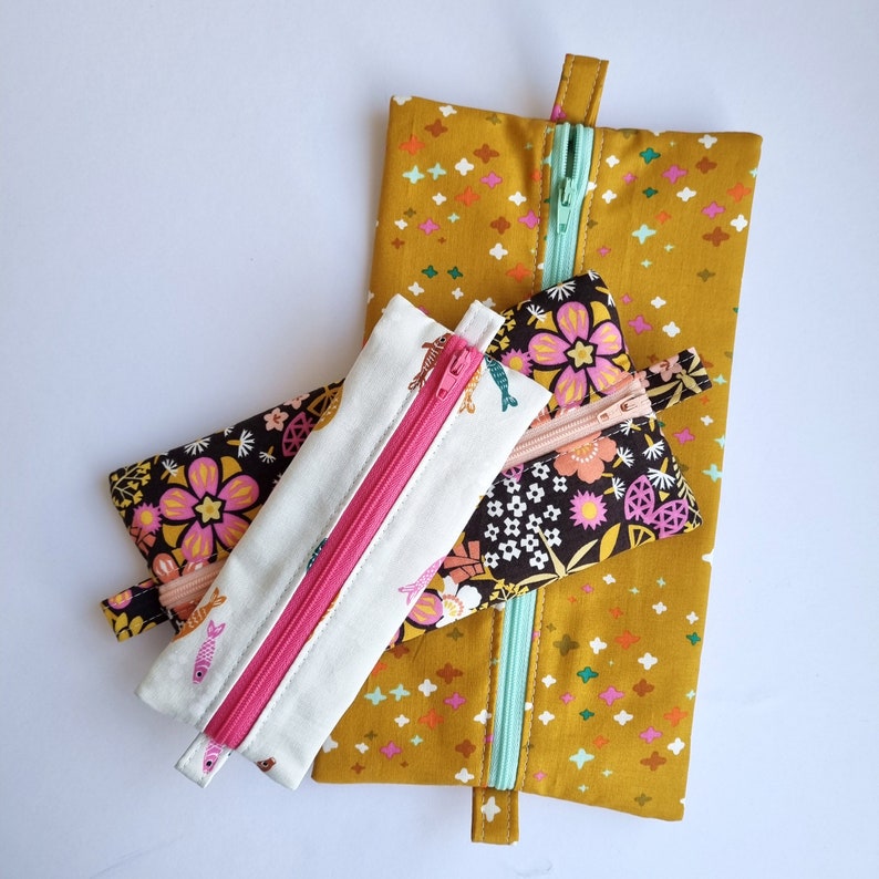 May include: Three zippered pouches in different sizes and patterns. The largest pouch is yellow with white polka dots. The medium pouch is black with pink and white flowers. The smallest pouch is white with pink fish and a pink zipper.